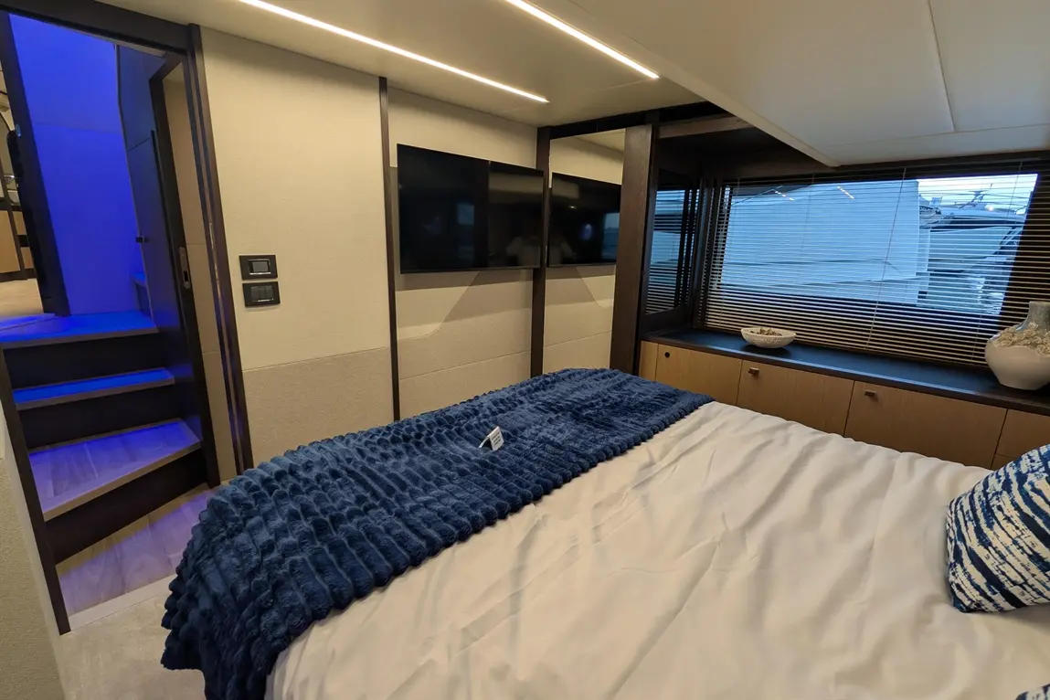 Modern bedroom featuring a blue blanket, two wall-mounted TVs, and a view of stairs with blue lighting. Window with blinds.